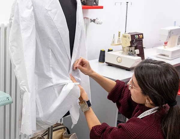 Studentessa spilla una tela di prova su un manichino da sartoria, con macchine da cucire e taglia e cuci sullo sfondo